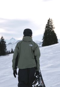 Personne portant une veste verte et un casque noir, portant un snowboard, debout sur la neige avec des pins et des montagnes en arrière-plan.