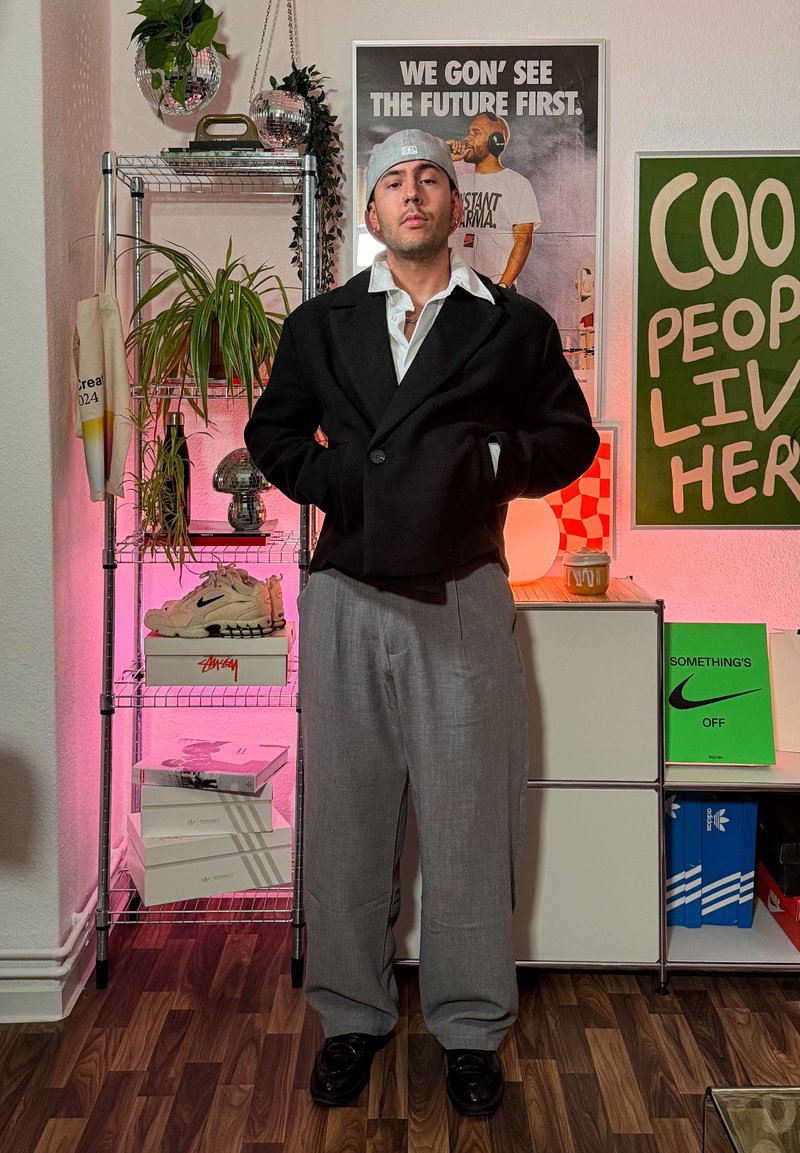 Black wool blazer, white shirt, light gray pleated trousers, black shoes. Accessories: gray beanie. Background includes plants and colorful posters.