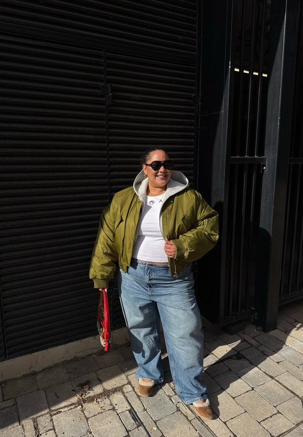 Una mujer sonriente con gafas de sol, una chaqueta bomber de color verde oliva y vaqueros de pernera ancha está de pie al sol, sosteniendo un paraguas rojo plegado junto a una verja metálica negra.