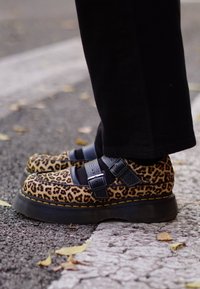 Kājām leoparddrukas platformas apavos ar melnām siksnām, stāvot uz asfalta pie saplaisājušas baltas līnijas ar izkaisītiem lapu gabaliņiem.