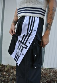 Μαύρο αθλητικό παντελόνι με λευκές ρίγες, που διαθέτει λογότυπο Adidas, τσέπες με φερμουάρ και κουμπιά στα πλάγια. Πλεχτό τοπ με ναυτικές ρίγες.