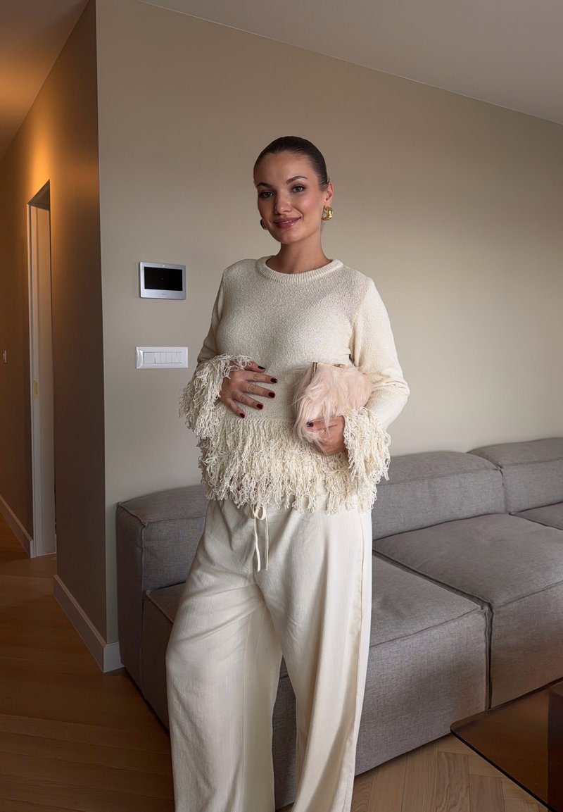 Une femme vêtue d’un pull crème à franges et d’un pantalon à jambes larges se tient près d’un canapé, un sac pochette rose pâle à la main.