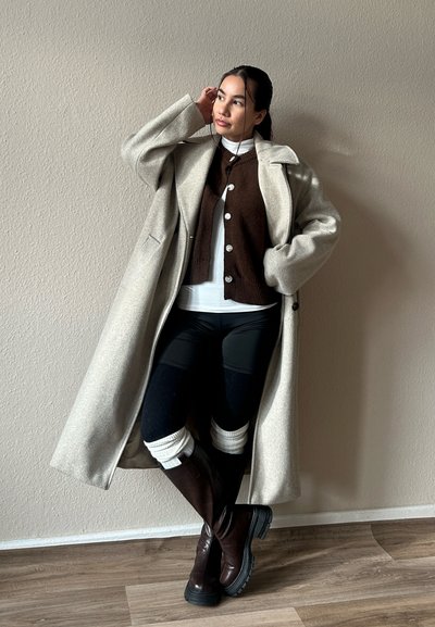 Mujer con un abrigo largo beige, suéter marrón con botones, legging negros, calentadores de piernas blancos y botas marrón oscuro, de pie contra una pared beige.