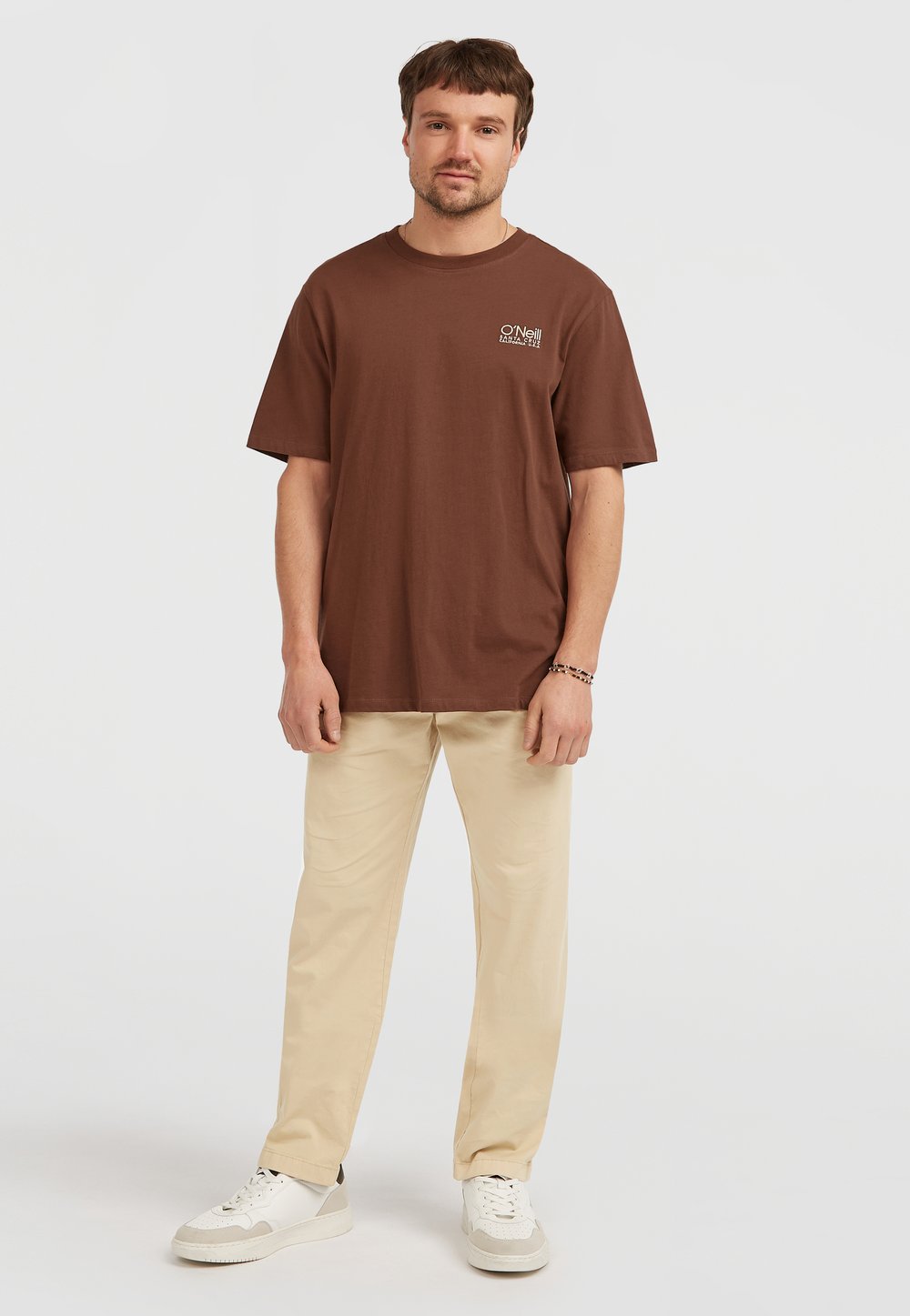 Brun bomulds t-shirt med korte ærmer og rund halsudskæring, parret med beige bukser og hvide sneakers. Enkel, casual design.