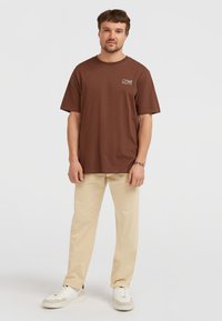 Brun bomulds t-shirt med korte ærmer og rund halsudskæring, parret med beige bukser og hvide sneakers. Enkel, casual design.