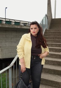 Žena s dugom tamnom kosom u žutom sakou, crnoj majici i trapericama stoji pored betonskih stepenica držeći crnu torbicu.