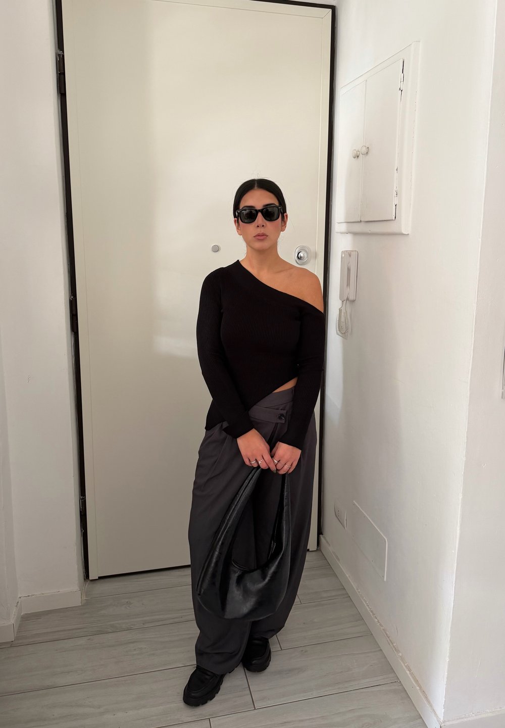 Haut noir à manches longues côtelé, pantalon gris à jambes larges, et lunettes de soleil noires. Tenant un sac noir texturé, debout devant une porte blanche.