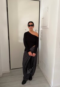 Haut noir à manches longues côtelé, pantalon gris à jambes larges, et lunettes de soleil noires. Tenant un sac noir texturé, debout devant une porte blanche.