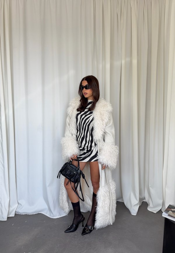 Mujer con abrigo largo blanco con ribete de piel, vestido mini con estampado de cebra, bolso negro, calcetines negros transparentes y tacones negros puntiagudos, de pie frente a cortinas blancas.