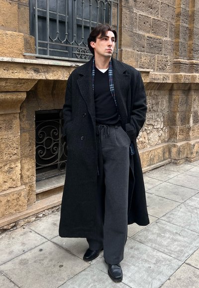 Hombre de pie en la acera vestido con un abrigo largo negro, suéter negro, pantalones anchos grises y zapatos negros, con las manos en los bolsillos cerca de un edificio de piedra.