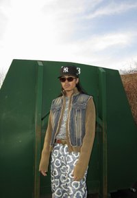 Gilet en denim avec des manches côtelées, t-shirt graphique gris, boucle de ceinture en cœur argent, et pantalon à motifs bleu et blanc. Lunettes de soleil marron et casquette.