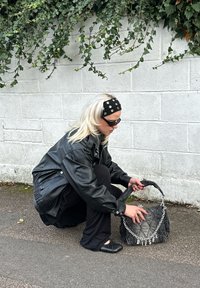 Prešívaná čierna kabelka so strieborným reťazovým detailom a logom "DIESEL". Mäkký materiál, zaoblený tvar, držaná osobou v čiernej koženej bunde.