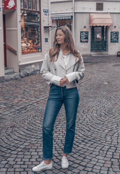 Abrigo tipo gabardina gris claro sobre una camisa blanca, combinado con jeans azules y zapatillas blancas. De pie en una calle empedrada, sonriendo suavemente.