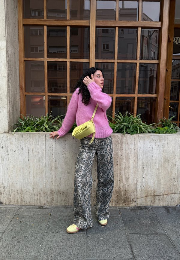 Femme en pull rose, pantalon imprimé serpent et sac jaune, debout sur le trottoir de la ville à côté d'un bac à fleurs en pierre avec des plantes vertes et une grande fenêtre en bois.