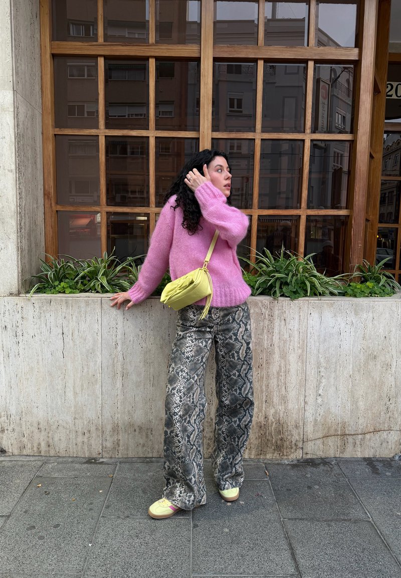 Femme en pull rose, pantalon à imprimé serpent et sac jaune, debout sur le trottoir de la ville à côté d'une jardinière en pierre avec des plantes vertes et une grande fenêtre en bois.