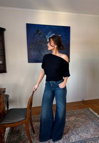 Femme aux cheveux bruns portant un haut noir à épaules dénudées et un jean pattes d'éléphant, tenant une chaise en bois dans une pièce avec une peinture abstraite bleue.