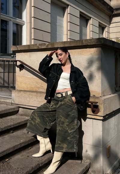 Chaqueta de mezclilla negra, top corto blanco, pantalones cargo anchos de camuflaje y botas de tacón blancas. Al aire libre, frente a una pared de piedra clara.