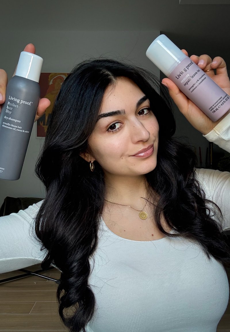 Femme aux longs cheveux bruns tenant deux produits de soins capillaires Living Proof, un shampooing sec et un soin sans rinçage, souriante à l’intérieur.