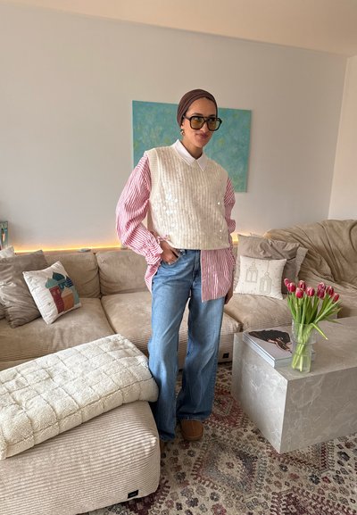 Mujer con gafas, camisa de rayas rosas, suéter sin mangas blanco, jeans azules, de pie en una sala de estar con un sofá beige y tulipanes sobre una mesa de centro.