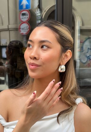 Pendientes de afirmación plateados con una superficie lisa y reflectante. Las uñas bien cuidadas tienen una base nude con diseños de estrellas en color burdeos. Blusa blanca sin hombros.