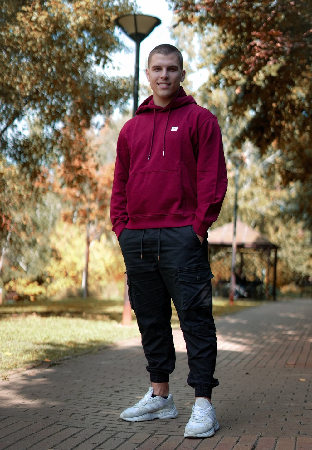 Bordó kapucnis pulóver zsinórral, fekete cargo nadrág zsebekkel és fehér sportcipő. Külső környezet fák és ösvény a háttérben.