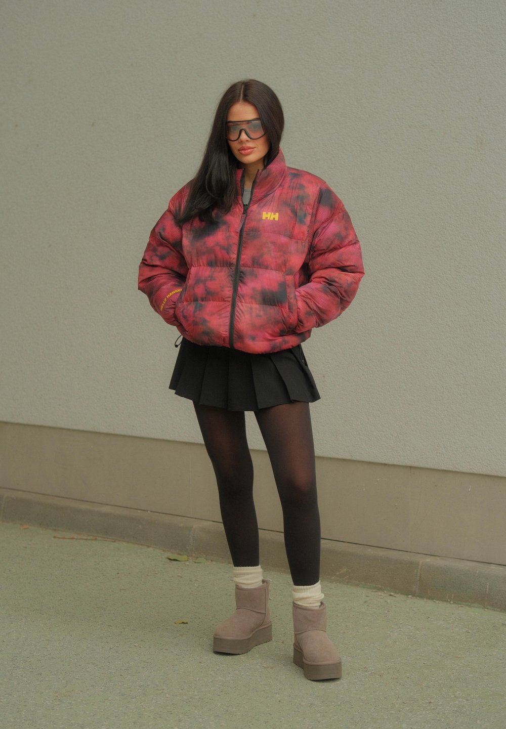 Κόκκινο και μαύρο καπιτονέ μπουφάν με σχέδιο και φερμουάρ, συνδυασμένο με μαύρη πλισέ φούστα, μαύρες καλσόν και μπεζ μπότα μέχρι τον αστράγαλο.