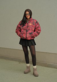 Κόκκινο και μαύρο καπιτονέ μπουφάν με σχέδιο και φερμουάρ, συνδυασμένο με μαύρη πλισέ φούστα, μαύρες καλσόν και μπεζ μπότα μέχρι τον αστράγαλο.