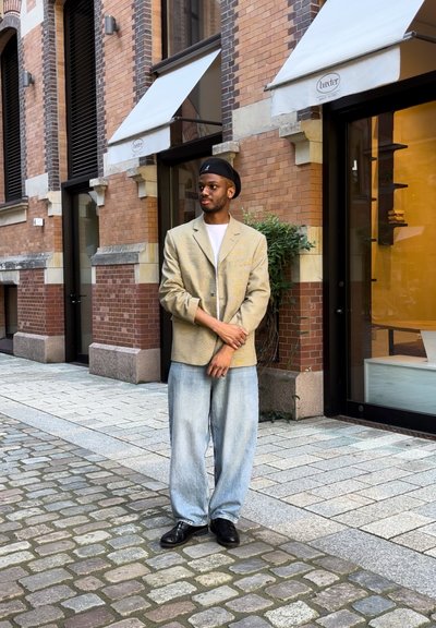 Blazer estampado beige, camiseta blanca y jeans azules holgados con zapatos negros, de pie en un camino de adoquines cerca de una pared de ladrillo.