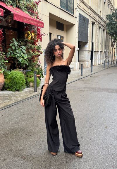 Mujer joven con cabello rizado, vestida con un top negro sin tirantes y pantalones anchos, de pie en la calle cerca de un restaurante italiano con plantas y un toldo rojo.
