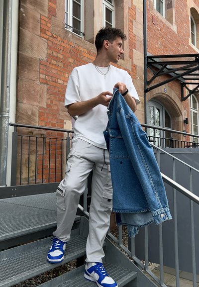 El atuendo informal incluye una camiseta blanca oversize, pantalones cargo grises y zapatillas azul y blancas, con una chaqueta de mezclilla colgada del brazo.