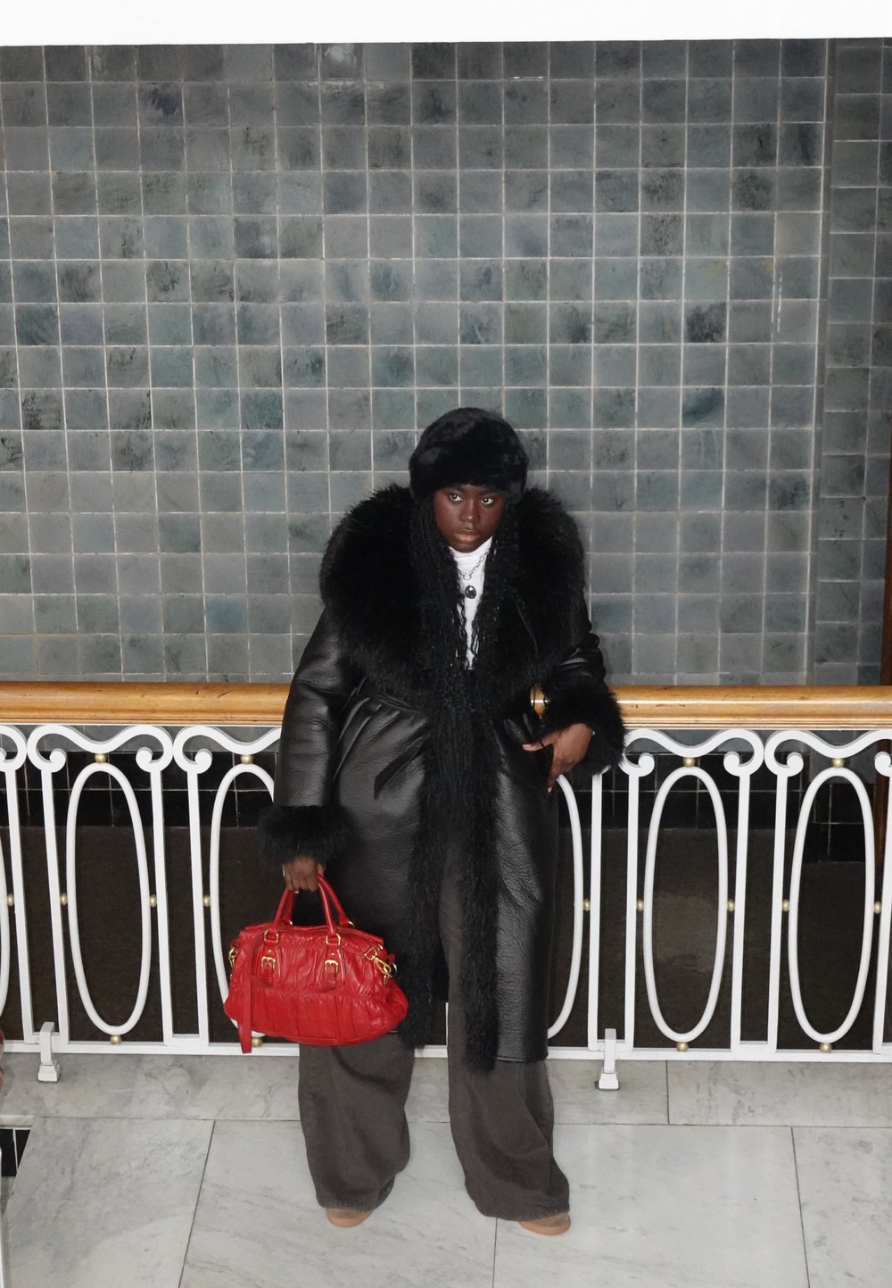 Manteau en cuir noir avec bordure en fausse fourrure, associé à un pantalon gris. Un sac à main rouge apporte une touche de couleur. L'arrière-plan présente un mur carrelé et une rampe.