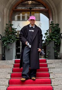 Melna garā mētelis virs grafiskā T-krekla, redzams ruļļa krekla apakšdalis. Tumši džinsi, brūni apavi, tur rokā lietussargu. Sarkanas paklājs un zaļumi fona.