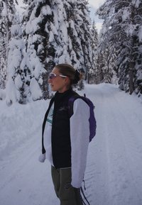 Black puffy gilet over a grey sweatshirt, green logo, olive trousers, purple backpack, snow-covered trees in the background, bright light.