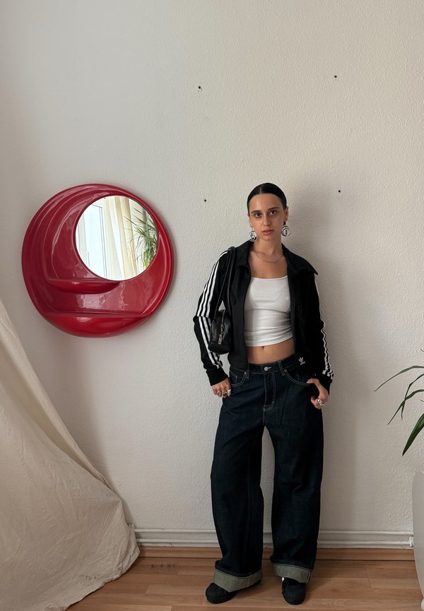 Fekete cipzáras dzseki fehér csíkkal, fehér crop top, laza sötét farmer, felhajtott szárakkal, és fekete cipő. Piros kör alakú tükör és növény a háttérben.