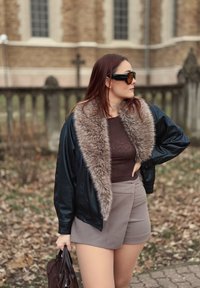 Černá kožená bomber bunda s umělým kožešinovým límcem, hnědý přiléhavý top, béžové kraťasy s vysokým pasem a tmavá kabelka, doplněné slunečními brýlemi.