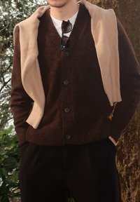 Άτομο που φοράει καφέ ζακέτα με κουμπιά και μπεζ πουλόβερ περασμένο πάνω από τους ώμους, με γυαλιά ηλίου κολλημένα στη ζακέτα, που στέκεται δίπλα σε δέντρο.