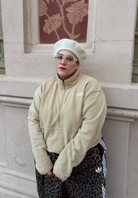 Personne portant une veste polaire beige, un béret blanc avec un logo, de grandes lunettes, un anneau au nez et un pantalon Adidas à motif léopard, debout contre un mur beige avec une fresque florale.