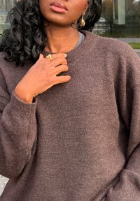 Rudas megztinis laisvo silueto, apvalia iškirpte ir tekstūruota medžiaga. Ant rankos matomas ryškus auksinis žiedas, taip pat matomi dideli žiediniai auskarai.