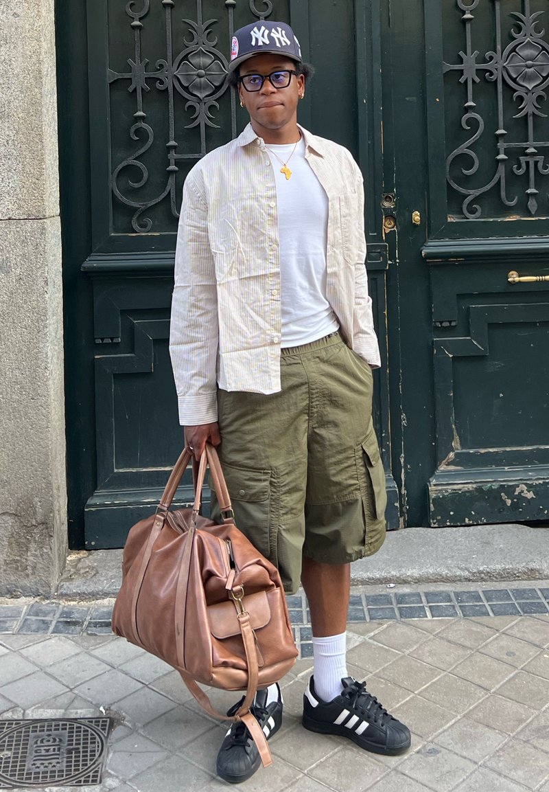 Tenue décontractée comprenant une chemise légère à rayures beige, un t-shirt blanc, un short cargo vert olive, des baskets noires et un sac de voyage en cuir marron.