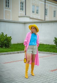 Ženska v rumeni klobuki, rumenih čevljih, dolgem roza kardiganu, modri srajci in bež kratkih hlačah, drži rumeno torbico in stoji na tlakovanem poti.