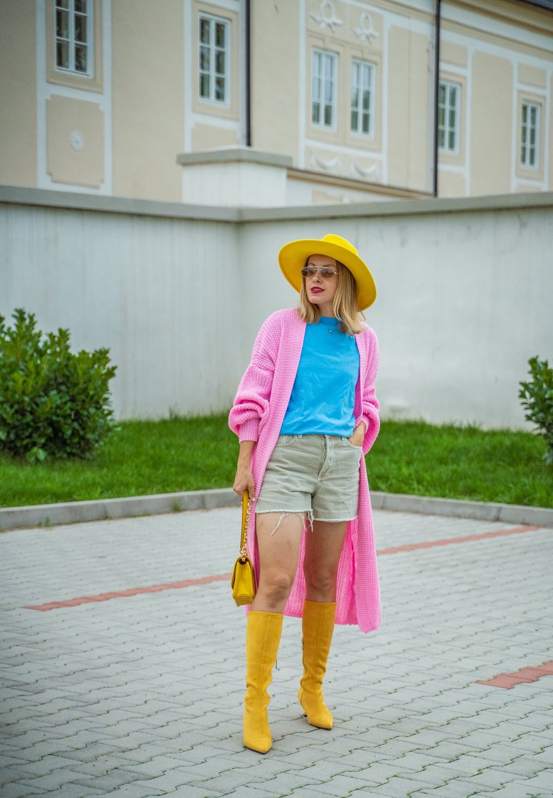 Mulher a usar um chapéu amarelo, botas amarelas, um cardigan longo rosa, uma camisa azul e calções beges, segurando uma mala amarela, de pé num caminho pavimentado.
