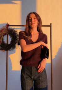 Femme portant un pull à manches courtes violet foncé et un jean, tenant une pochette noire, debout à côté d'un présentoir en bois avec une couronne décorative.