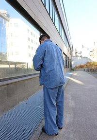 Gaiši zila džinsu jaka un plata kāju džinsi. Jakai ir brīvs piegriezums un redzamā šuvšana. Betons ar stikla atspulgajiem.