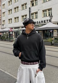 Jeune personne portant des lunettes et un sweat à capuche noir debout dans une rue de la ville, tenant des clés, avec des places assises en plein air et des fenêtres de bâtiment en arrière-plan.