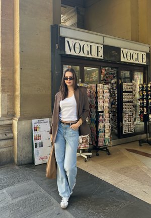 Mujer con gafas de sol, blazer marrón, camiseta blanca y jeans, llevando un bolso marrón, de pie frente a un kiosco de Vogue Italia con revistas y souvenirs.