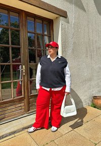 Μαύρος άκρυφος φορμαρισμένος πουλόβερ πάνω από λευκό πουκάμισο με κουμπιά, κόκκινα παντελόνια, κόκκινο καπέλο και λευκή τσάντα. Γκρίζα και λευκά αθλητικά παπούτσια. Ξύλινη πόρτα στο φόντο.