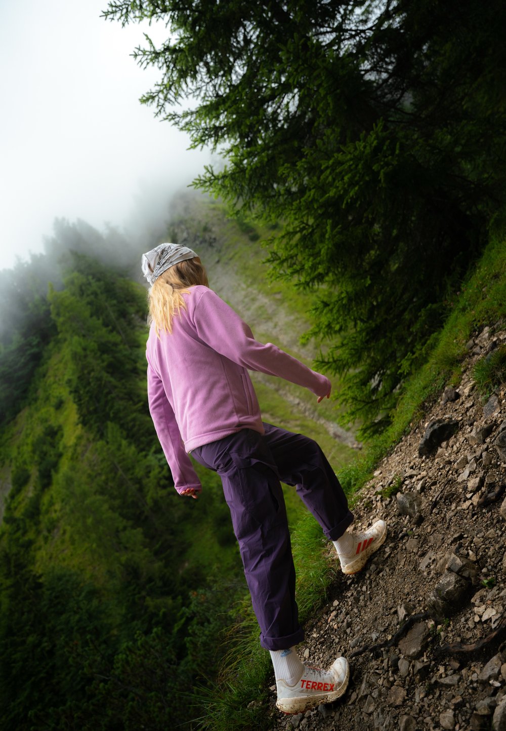 Wandelscène met een persoon in een roze fleecejack, paarse broek en witte TERREX-schoenen, die over een rocky pad tussen groene bladeren navigeert.