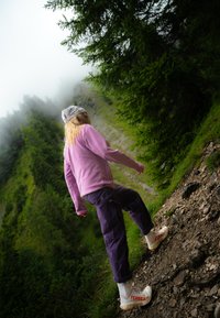 Wandelscène met een persoon in een roze fleecejack, paarse broek en witte TERREX-schoenen, die over een rocky pad tussen groene bladeren navigeert.