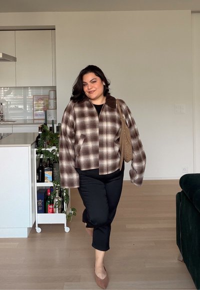 Mujer caminando en interiores con una chaqueta de cuadros marrón, pantalones negros, zapatos planos marrones y llevando un bolso de hombro color camel.