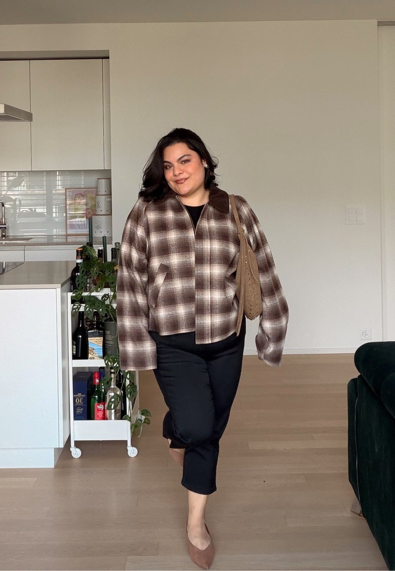 Femme marchant à l'intérieur, portant une veste à carreaux marron, un pantalon noir, des ballerines marron, et portant un sac à main couleur beige.